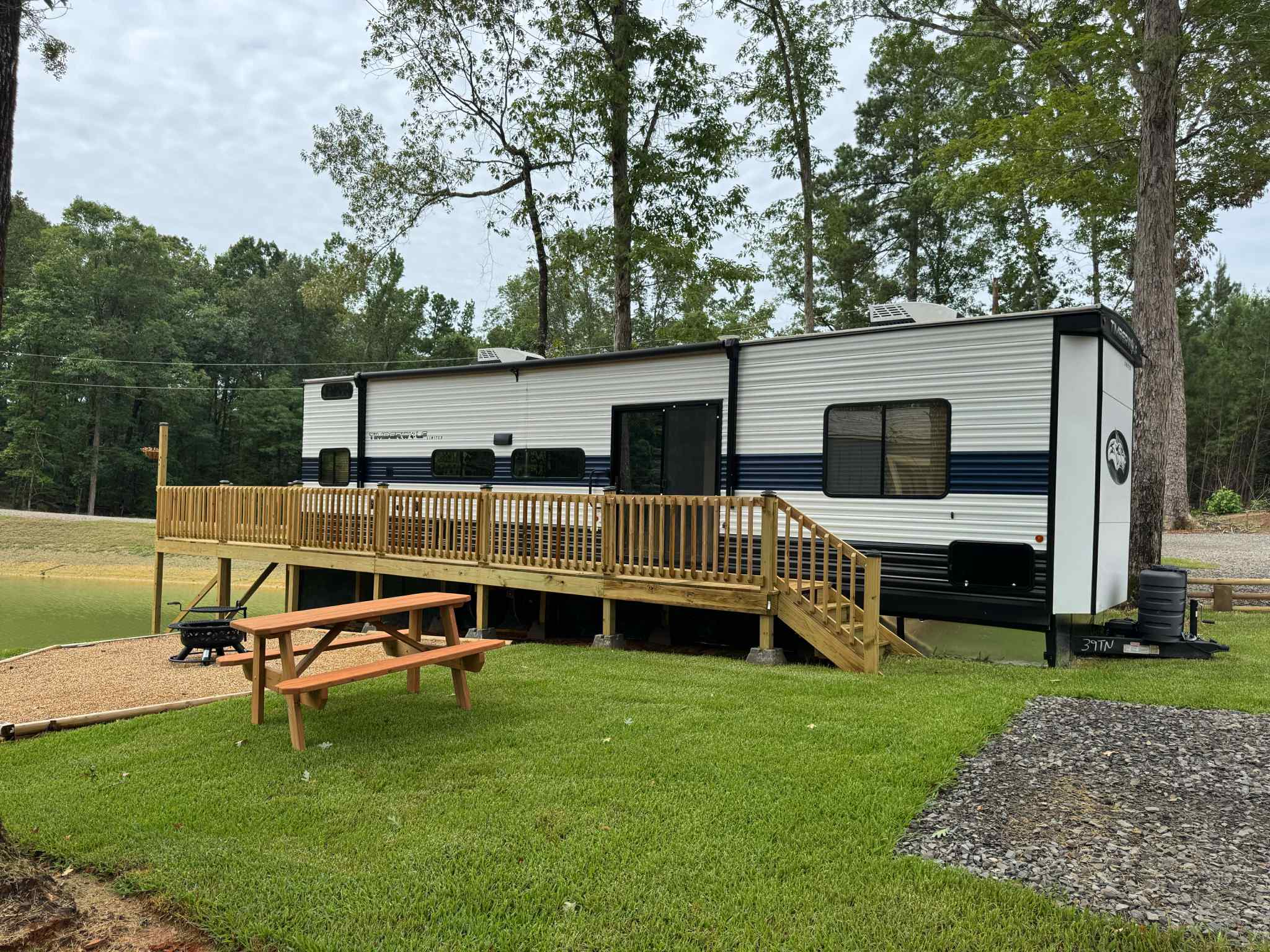 A parked RV with a wooden deck beside a pond, surrounded by grass and trees, with a picnic table and gravel area in the foreground.