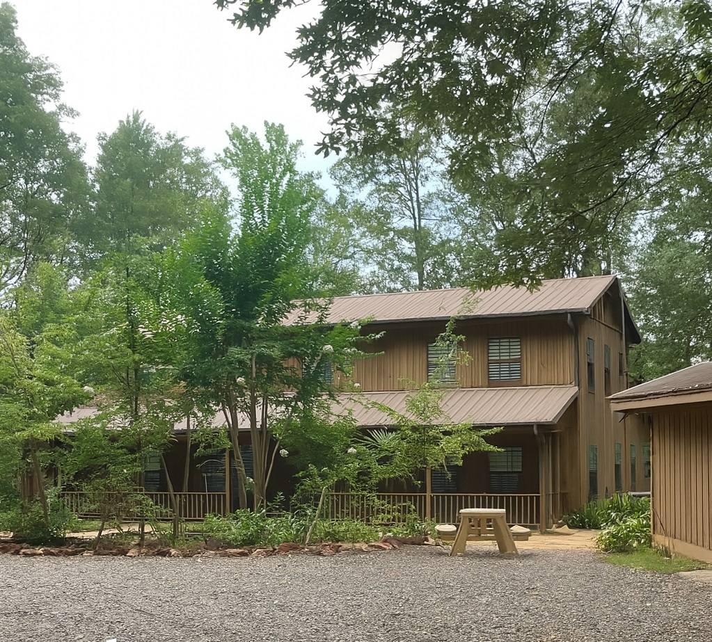 A two-story brown wooden building with a metal roof sits among trees, with a gravel path and bench in the foreground.